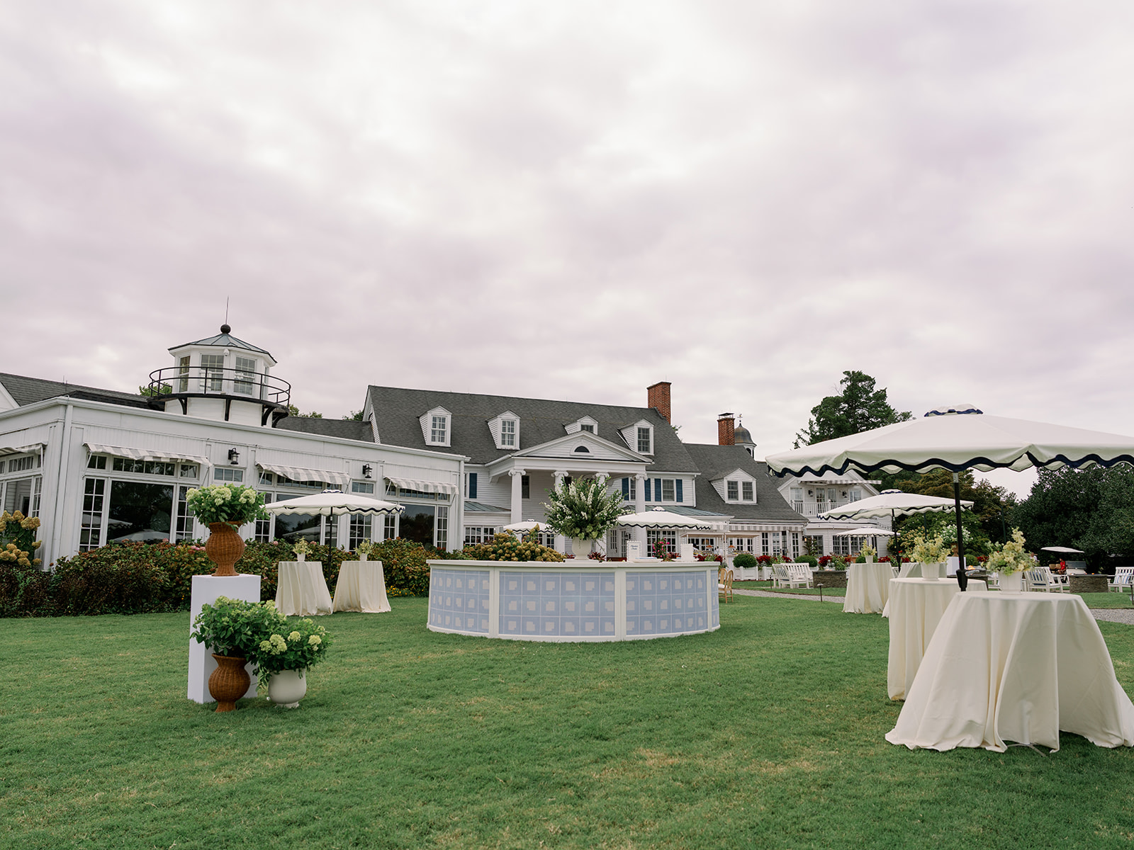 A picturesque outdoor setup with tables, umbrellas, and greenery in front of a large building with multiple windows and a lighthouse-like structure.