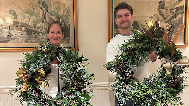 Two people are holding decorative wreaths made of greenery, pinecones, and ribbons. Duck paintings are visible in the background.