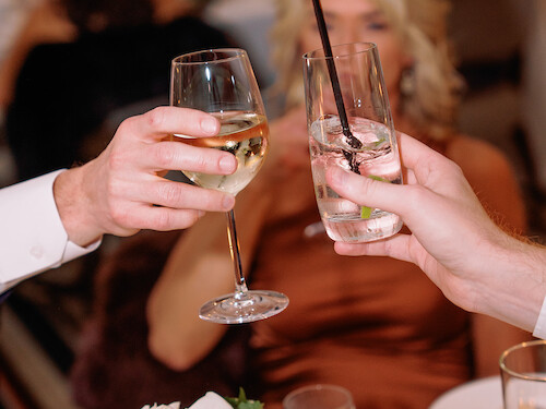 Two people clinking glasses at a formal event with a blurred background and floral centerpiece in view.