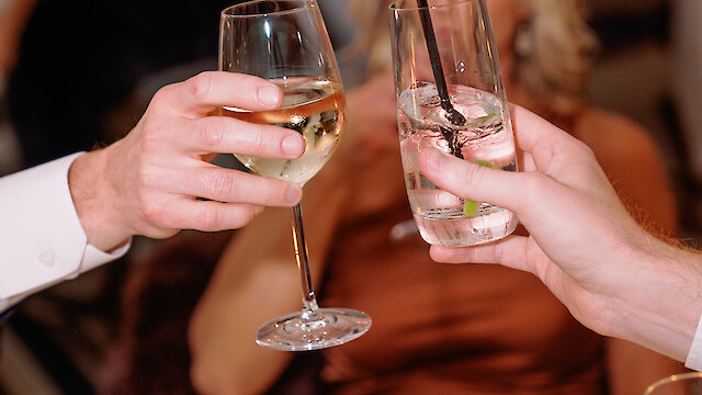 Two people clinking glasses at a formal event with a blurred background and floral centerpiece in view.